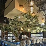 Blake’s Oyster Bar interior in Atlanta featuring a custom tree bar, oyster-back bar stools and statement lighting, designed by Contemporary LUX. Custom restaurant bar stools featuring intricate, double-layered metal backrests shaped into the oyster-inspired restaurant logo, surrounding a dramatic tree installation in an Atlanta oyster bar.