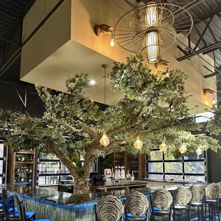 Blake’s Oyster Bar interior in Atlanta featuring a custom tree bar, oyster-back bar stools and statement lighting, designed by Contemporary LUX. Custom restaurant bar stools featuring intricate, double-layered metal backrests shaped into the oyster-inspired restaurant logo, surrounding a dramatic tree installation in an Atlanta oyster bar.