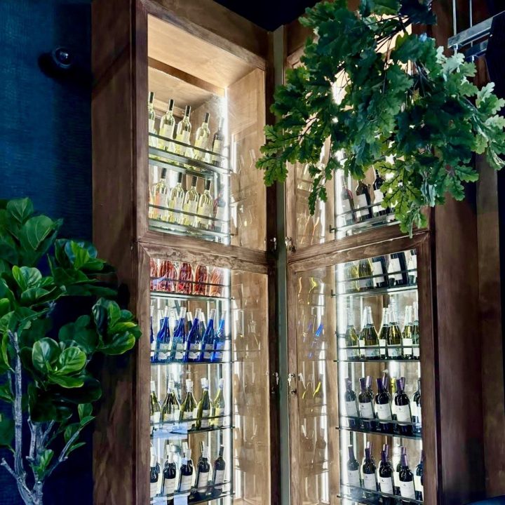 Custom floor-to-ceiling wood wine display cabinets featuring glass doors, illuminated glass shelving, and curated wine bottles, framed by lush greenery in a contemporary Atlanta restaurant.