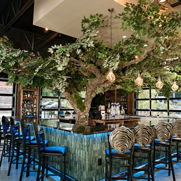 Custom bar stools featuring intricate double-layered metal backrests incorporating the restaurant's oyster-shaped logo, positioned along a blue-illuminated bar beneath a dramatic indoor tree installation.