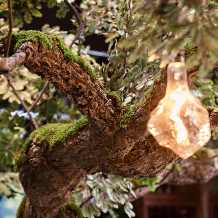 Close-up of a moss-covered tree installation with textured bark, lush foliage, and a glowing sculptural glass pendant in a New Orleans-inspired restaurant interior.