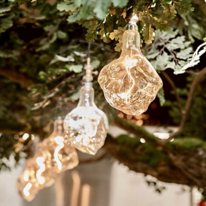 Close-up of sculptural, textured glass pendant lights suspended among the branches of an indoor tree installation, casting a warm glow in a New Orleans-inspired restaurant interior.